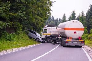 A car is crashed into the front of a tanker truck on a curved rural road surrounded by trees. Debris is scattered around the accident scene.