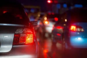 Cars in heavy traffic at night with red brake lights illuminated and wet road surfaces reflecting the lights.