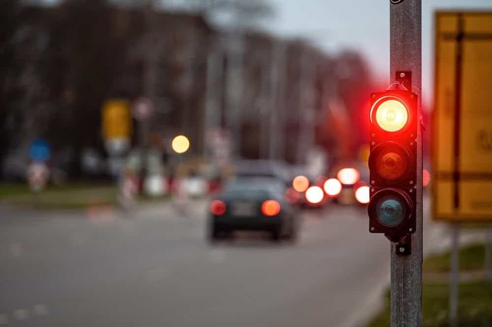 A road with a red traffic light in the foreground. Cars are blurred in motion in the background on a cloudy day.