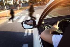 View from a car of two pedestrians crossing a road in a crosswalk on a sunny day. A driver's hand is visible on the steering wheel.