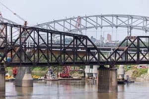 Steel bridges under construction over a river, with cranes and city skyline in the background.