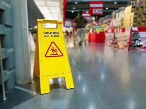 Yellow "Caution Wet Floor" sign on a glossy store floor with shelves of products and a person walking in the background.
