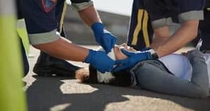 Two paramedics assist a person lying on the ground, securing a bandage on their head. The responders wear uniforms and blue gloves.
