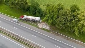 Aerial view of a road accident with a red truck overturned on the side, debris scattered, and a car parked nearby on a highway surrounded by trees and grass.