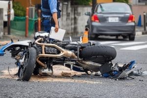 A motorcycle lies on its side on a road with scattered debris around it, while a person in a blue uniform stands nearby holding a document. A car is parked in the background.