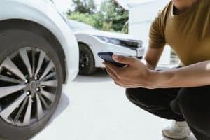 Person crouching near a white car, holding a smartphone, observing the vehicle. Another car is parked in the background.