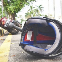 A motorcycle helmet lies on the ground with a fallen motorcycle in the background, next to a yellow line on a road. Trees and buildings are visible in the distance.