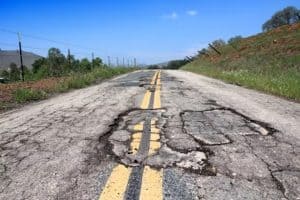 A rural road with yellow lines is heavily cracked and damaged with several large potholes. The background shows greenery and a clear sky.