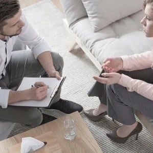 A man with a clipboard and pen listens to a woman sitting on a couch during a conversation, possibly in a counseling or meeting setting.