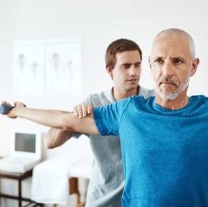 A man holds his arm out for an exercise while another person assists.