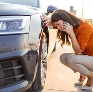 Woman crouches by a car with a flat tire, talking on her phone, appearing concerned.