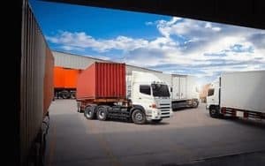 Trucks with shipping containers parked in an industrial loading area under a cloudy sky.
