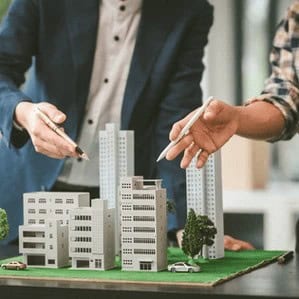 Two people discussing a miniature architectural model of a cityscape with buildings and trees on a table.