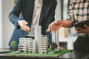 Two people discussing a detailed architectural model of a modern cityscape, which includes multiple buildings and greenery. One holds a tablet, while both point at different structures.