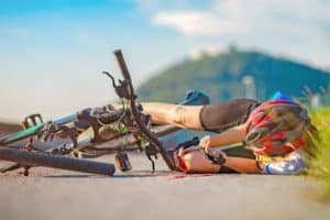 Cyclist wearing a helmet lies on the ground beside a fallen bike on a sunny day, with a hill in the background.