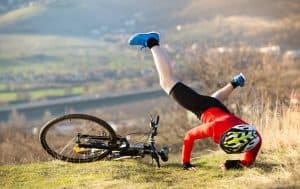 Cyclist in a red shirt and helmet falls headfirst beside a bike on grassy terrain, with a hilly landscape in the background.