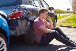 A man in a red shirt sits on the ground next to a damaged car, talking on a phone, with another vehicle nearby on a sunny road.
