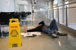 A person lying on the floor of a hallway next to scattered papers, a laptop, and a caution sign indicating a wet floor.