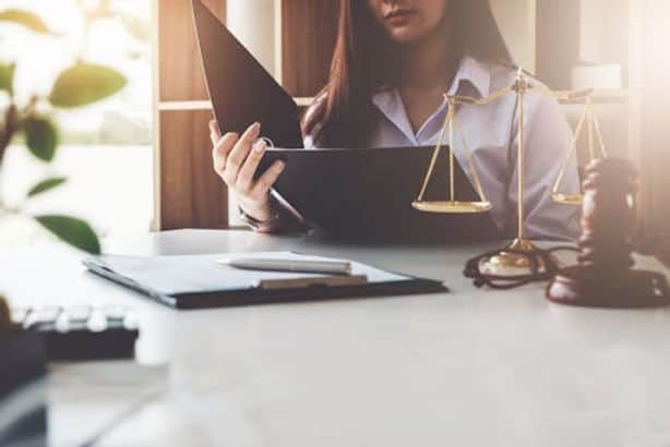 A woman reads a document at a desk with legal scales, a gavel, and a clipboard in a sunlit office.