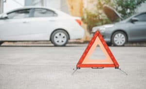 A red caution triangle placed on the road, with two parked cars in the background, one with the hood open.