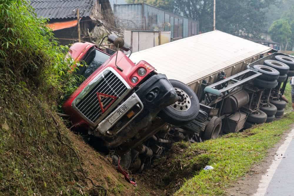 Overturned red truck on a grassy roadside, partially in a ditch, with its wheels in the air and a white cargo trailer attached.