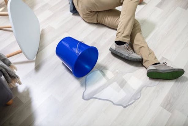 A person sitting on the floor next to a spilled blue bucket of water, with the water forming a puddle on the light wood floor. A chair is tipped over nearby.