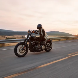 Rider on a black motorcycle travels down a highway at sunset, with hills and fields in the background.
