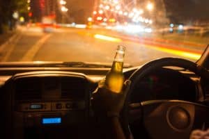 Person holding a beer bottle while driving at night, with blurred city lights visible through the windshield.