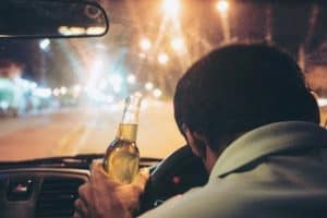 Person driving at night holding a bottle of alcohol close to their face with both hands on the wheel.