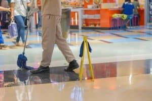 Person mopping a shiny floor in a mall with a caution sign nearby, food court and shoppers visible in the background.