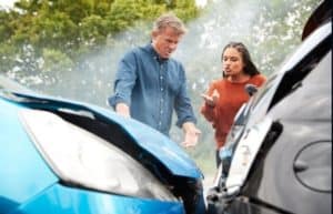 Two people stand near two cars with damaged front ends. The man gestures, looking frustrated, while the woman gestures at the damage. Smoke is visible in the background.