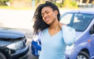 A woman in a blue sweater stands between two cars after an accident, holding her neck in discomfort.