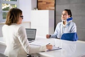 Two women in an office setting. One is wearing a neck brace and arm sling. They are seated at a table with documents and a laptop.