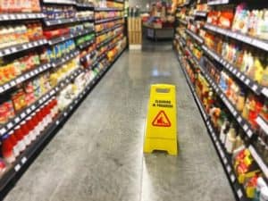A yellow "cleaning in progress" sign is placed in the center of a grocery store aisle with shelves filled with various products on both sides.