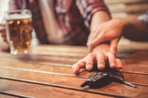 A person reaches for car keys on a wooden table as another hand stops them. A glass of beer is in the background.