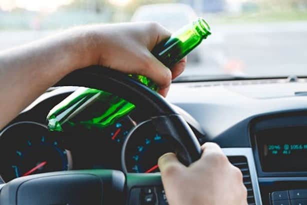 A person holds a green bottle and steers a car.