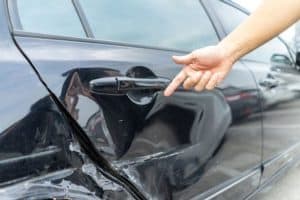A person points at a large dent and scratches on the side of a black car.