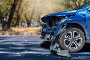 Front-left view of a blue car with significant damage, including a missing bumper and exposed internal parts, parked on a road with trees in the background.