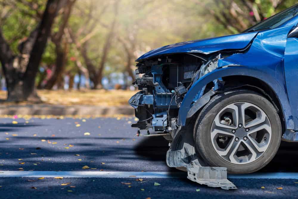 Front-left view of a blue car with significant damage, including a missing bumper and exposed internal parts, parked on a road with trees in the background.