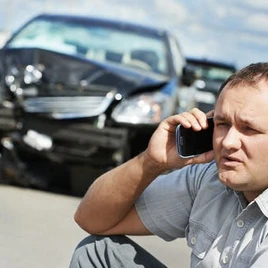 A man on a cellphone sits on a curb near a damaged car, possibly reporting an accident.