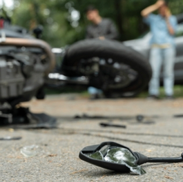 A fallen motorcycle on the road with shattered glass nearby. Two people stand in the background, one with hands on their head.
