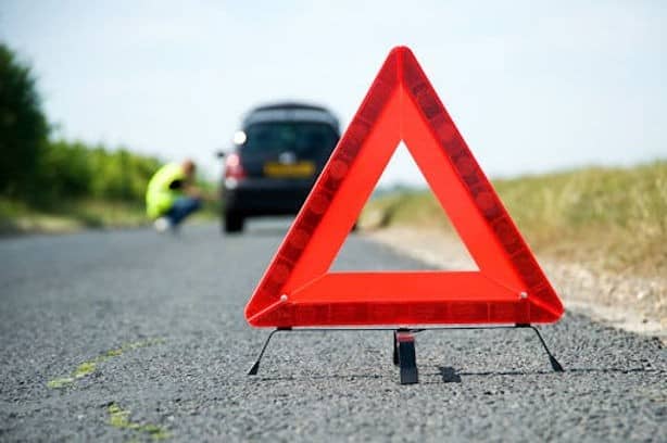 A red warning triangle on a road with a car and a person in a safety vest in the background.