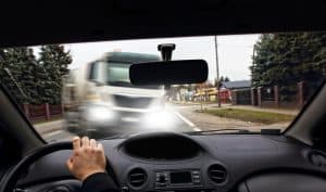 View from inside a car approaching an oncoming truck on a road. The driver's hand is on the steering wheel, and the street is lined with trees and buildings.