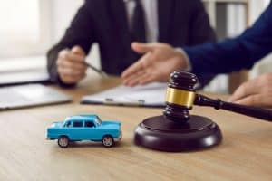 A toy car and gavel sit on a table in front of two people in suits, one gesturing and one holding a pen near a clipboard.