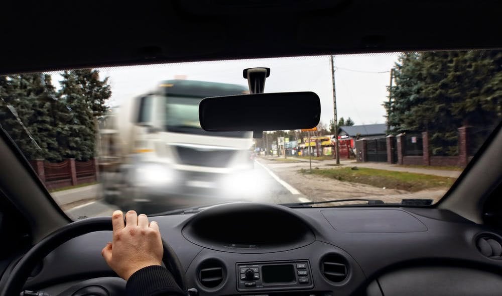 View from inside a car approaching an oncoming truck on a road. The driver's hand is on the steering wheel, and the street is lined with trees and buildings.