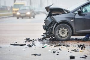 Severely damaged front of a black car after a collision, with debris scattered on a wet road. Other vehicles are visible in the background.
