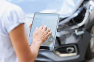 Person using a tablet in front of a damaged car, possibly documenting the damage or filing an insurance claim.