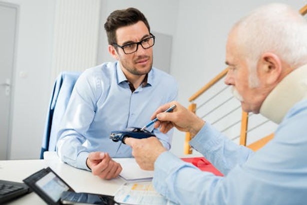 An older man wearing a neck brace discusses with a younger man across a table with documents and a pen.