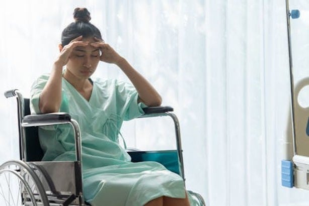 A woman in a hospital gown sits in a wheelchair with her hands on her forehead, near medical equipment.