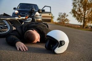 A person lies on the road near a fallen motorcycle and a white helmet, with an open car door visible in the background.